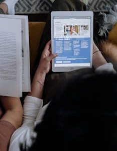 Overhead view of a person reading news on a digital tablet while holding an open book at home.